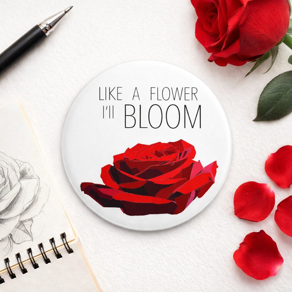 Round magnet with a red rose illustration and the quote “Like a flower I’ll bloom” on a styled desk with petals and sketchbook