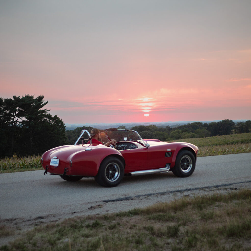 Red vintage convertible sports car driving on country road at sunset, scenic landscape with open fields and colorful sky.
