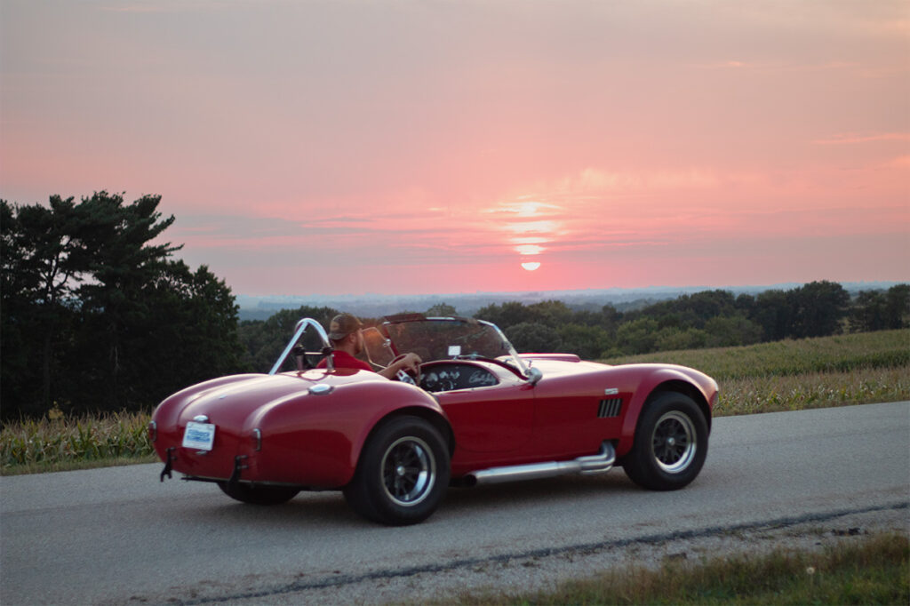 Red convertible driving on a rural road at sunset with a pink sky and open landscape