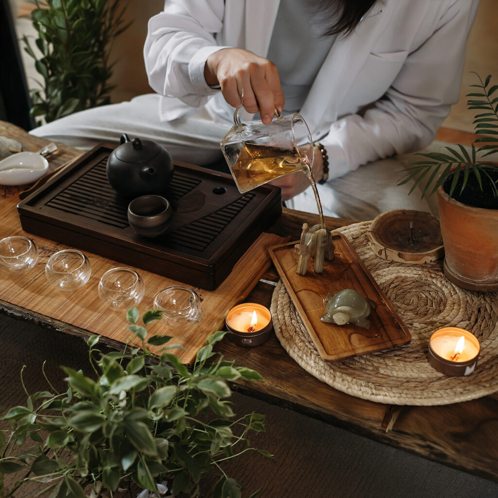 Traditional tea ceremony setup with person pouring tea from glass pitcher, featuring teapot, cups, candles, and wooden tea tray in a calm ritual setting.