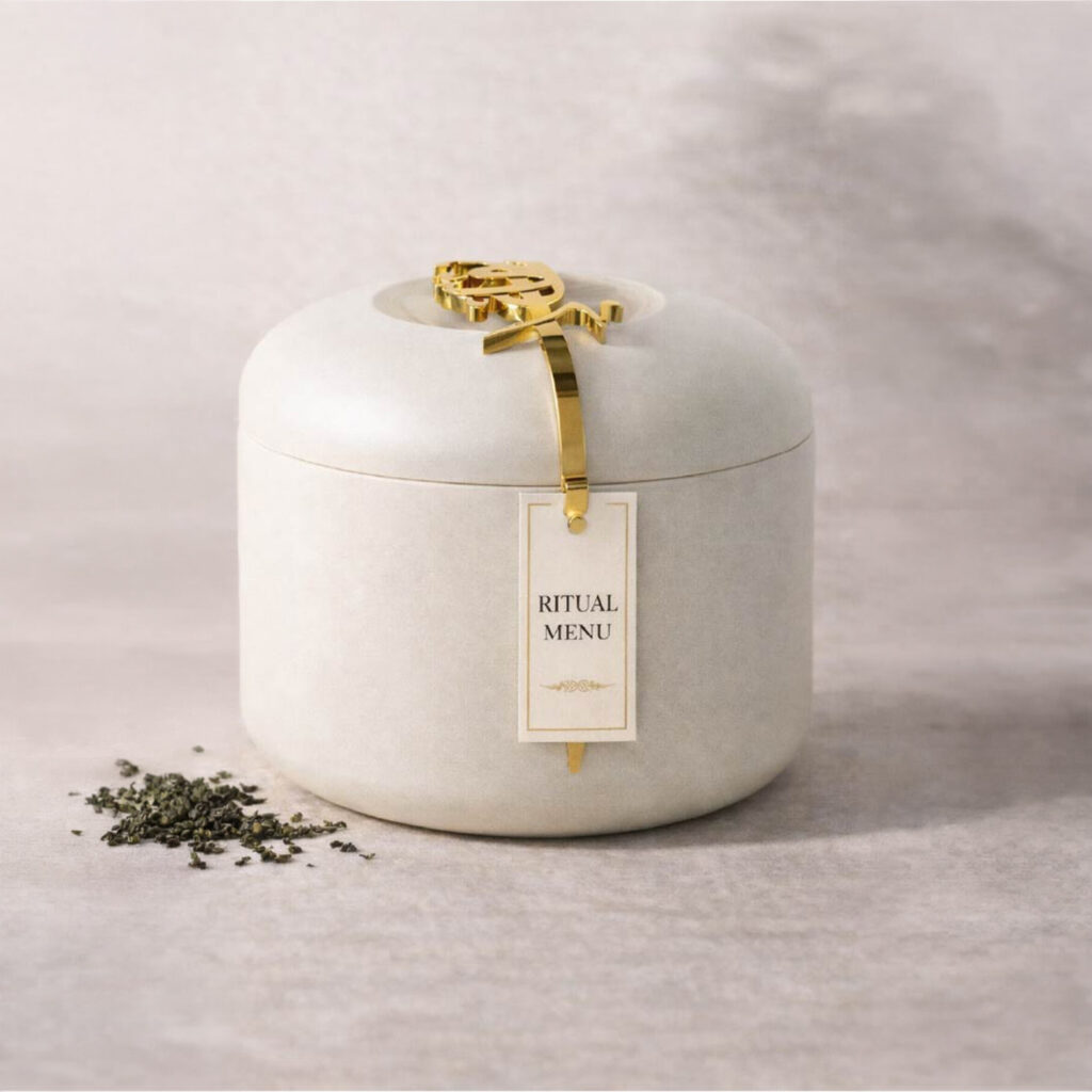 Luxury tea packaging container with gold clasp and ritual menu tag, minimal neutral design with loose tea leaves beside it.