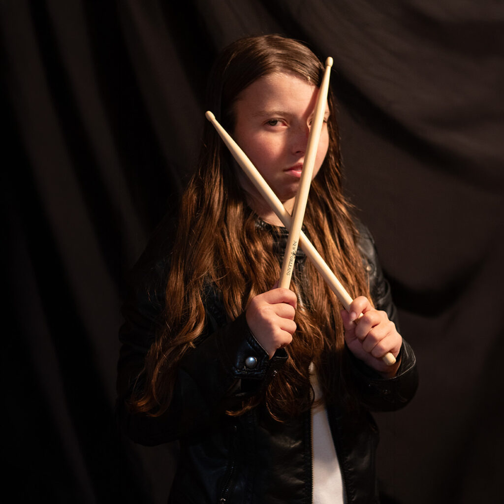 Dramatic portrait of girl holding drumsticks crossed in front of face, studio lighting with dark background and strong shadows.